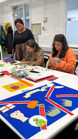 Two women at a table, crafting with paper while another woman is standing behind them, supervising.