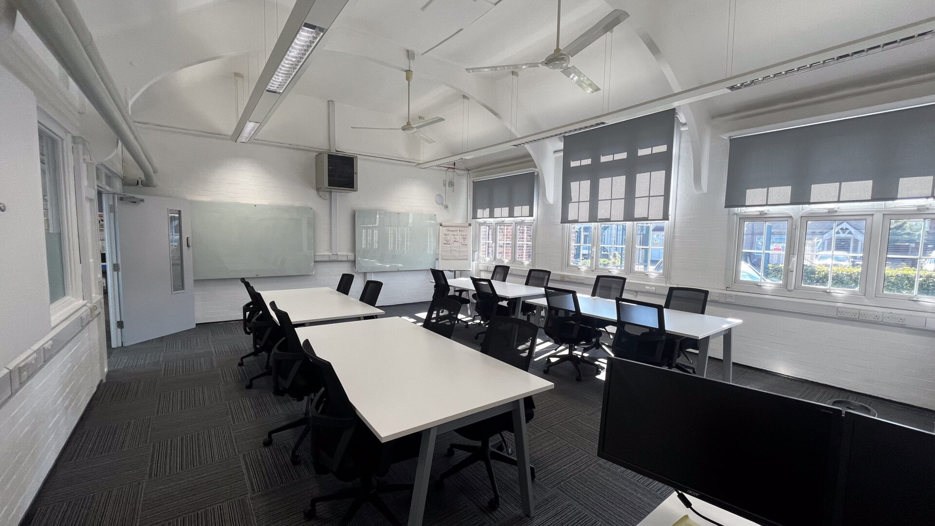 A room with white tables. black officer chairs and glass dry-wipe boards in the back