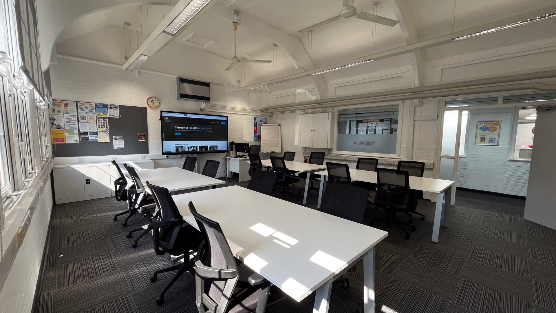 A room with white tables. black officer chairs and a large screen in the back