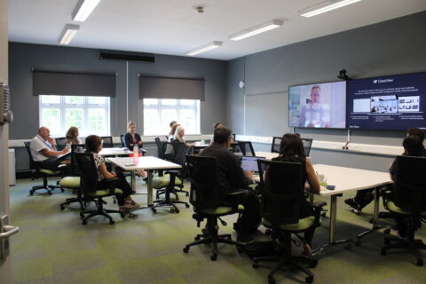 People sitting around desk looking at a screen where a man is shown