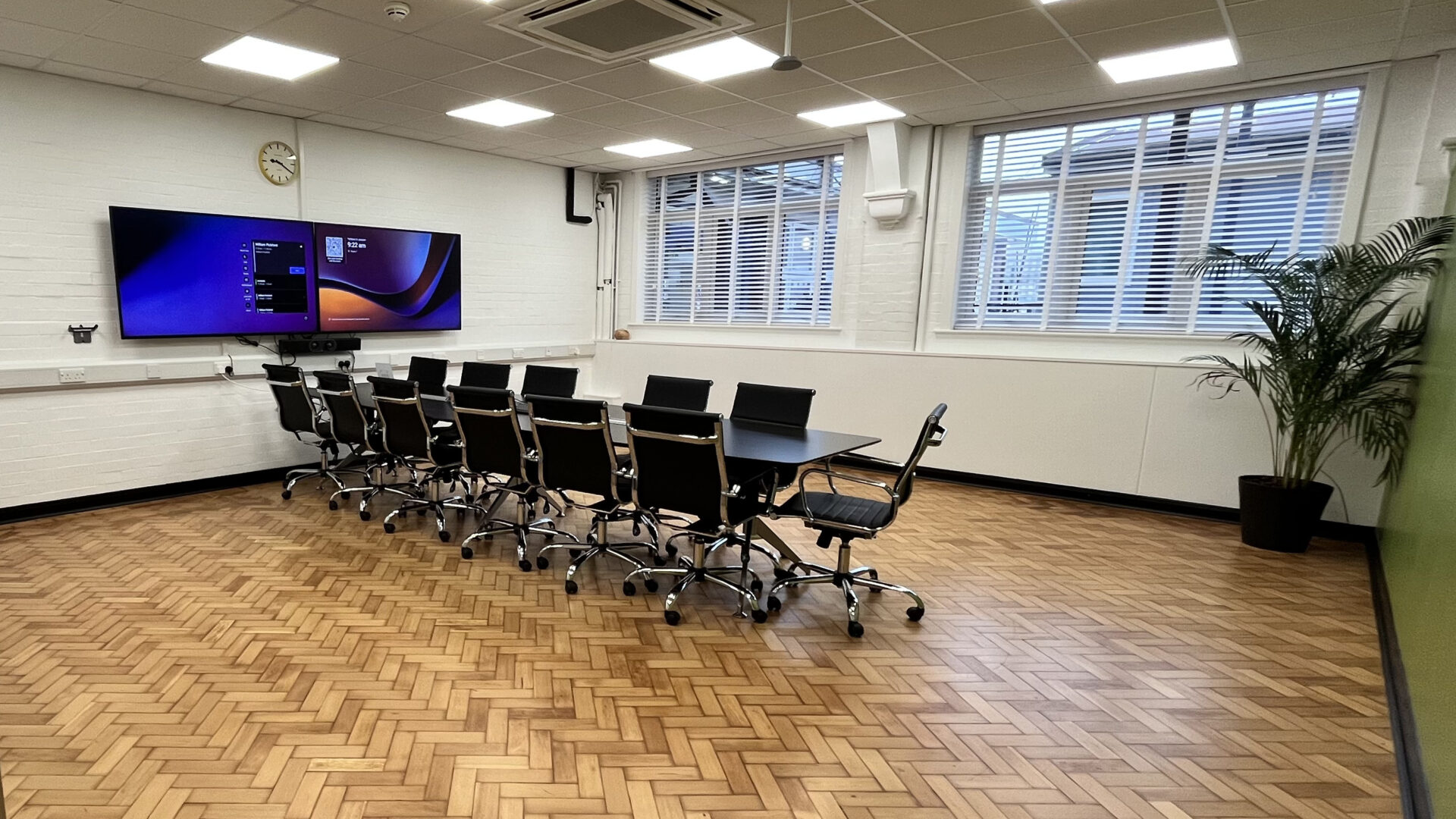 A meeting room with a large dark table and dark chairs in front of 2 screens