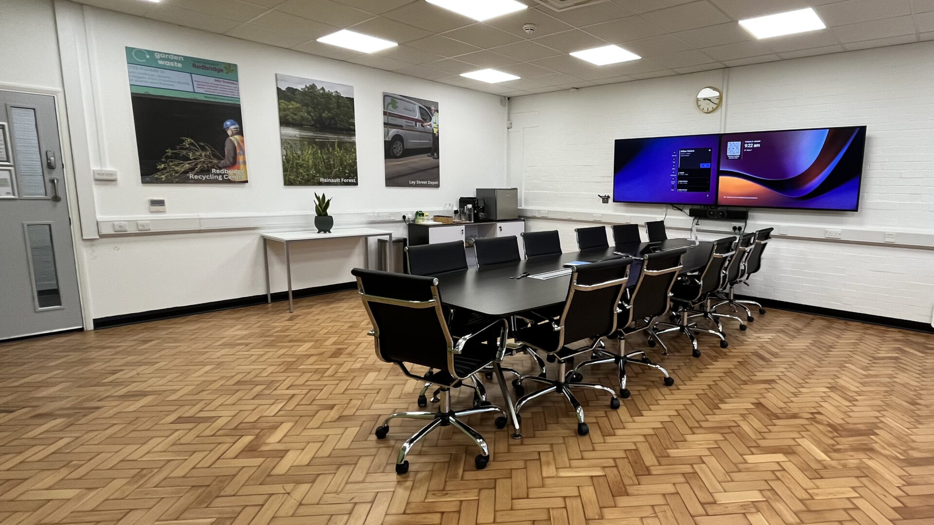 A meeting room with a large dark table and dark chairs in front of two screens and three pictures on the wall