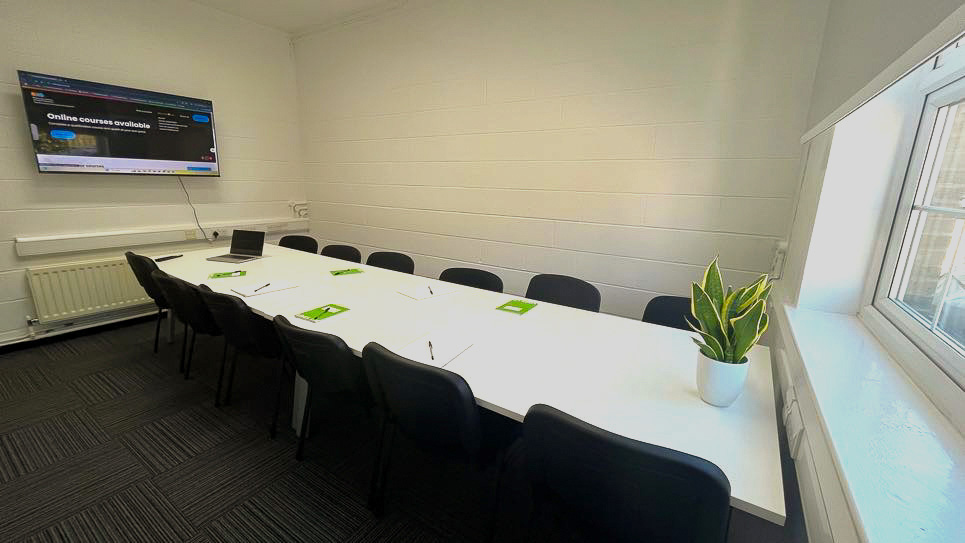 A long, white table with black chairs around it. A flipchart board sits on one end and a large screen on the other end.