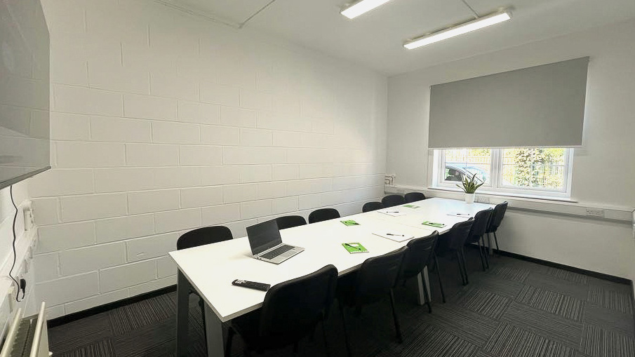 A long, white table with black chairs around it. A flipchart board sits on one end and a large screen on the other end.