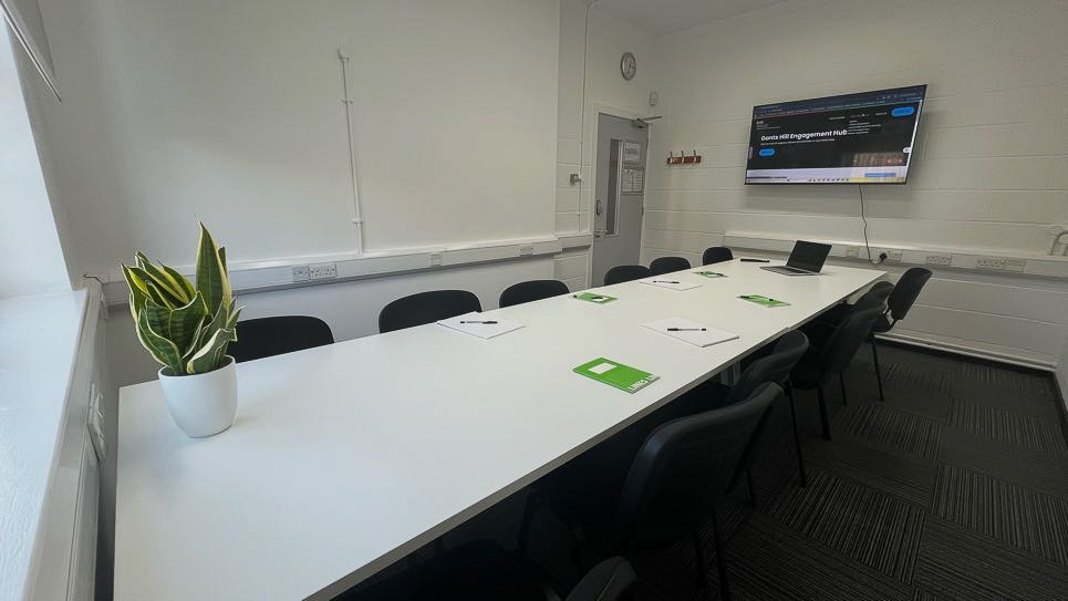 A long, white table with black chairs around it. A flipchart board sits on one end and a large screen on the other end.