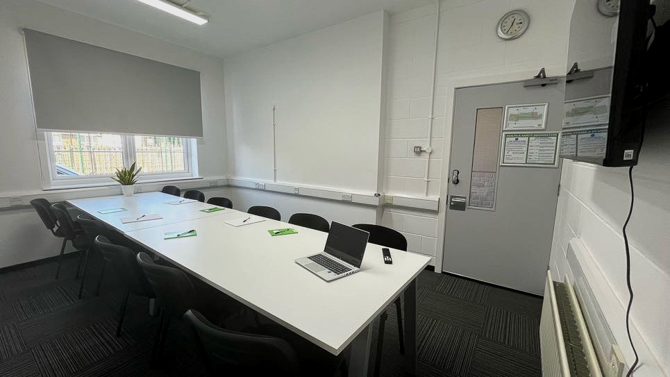 A long, white table with black chairs around it. A flipchart board sits on one end and a large screen on the other end.