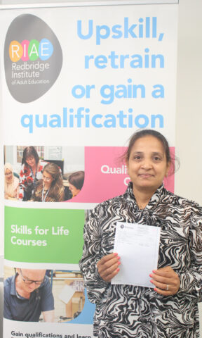 Learner standing in front of a Redbridge Institute banner holding a paper with their GCSE results